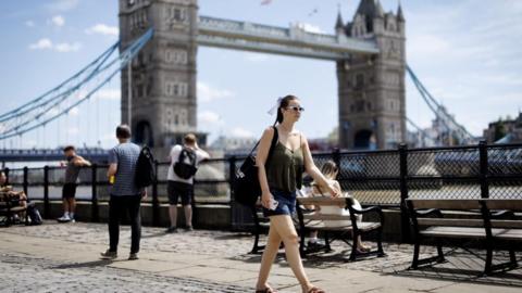 People near the River Thames during a warm day in London on 27 June 2025. A woman in shorts and a short sleeve top is at the centre of the image 