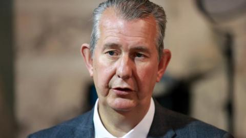 A head shot of Edwin Poots. He has grey hair and is wearing a grey blazer and white shirt.