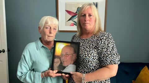A mother and daughter are standing in a living room holding a picture of their husband/dad.  The man in the picture is wearing a white shirt and is smiling.  He has a bald head and is wearing thin glasses on his face. He looks about 60-years old in the photograph.