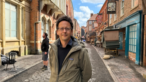 A slim man with dark-brown hair smiles as he stands in a cobbled street which has been dressed up to look like Victorian London with old signs, stalls and street furniture. He is wearing rectangular glasses and a green coat over a black jumper. Another man stands behind him looking towards a grand-looking building. 