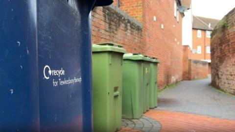 A photo showing a row of blue and green wheelie bins beside a red brick wall on a narrow street