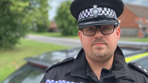 Ch Insp Nathan Shepherd looks at the camera, wearing a police cap and black jacket with the word POLICE on the front. Behind him is a neon yellow and blue police car.