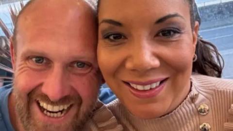 A man and woman are smiling at the camera in a selfie. She has her hair tied back and a beige ribbed jumper with frilled shoulders and gold buttons. He is wearing a blue tee shirt and has a bald head and ginger beard.