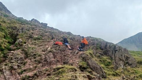 A picture of Will and Lyn at the site of the incident. Lyn is wearing a bright orange coat and black hat, while Will wears a cap, fleece and blue shorts. 