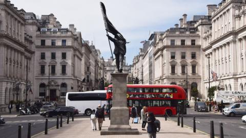 Statue of a man holding a flag in central London. A London bus and people walking around in the background
