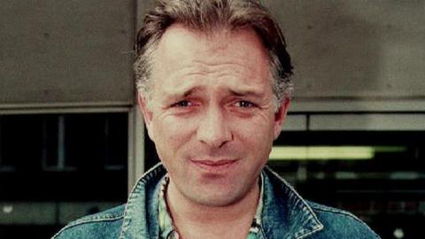 Rik Mayall looking directly towards the camera wearing a blue denim jacket. He is clean shaven and has brown swept back hair.