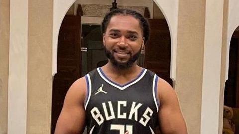 Person in a black Milwaukee Bucks jersey and shorts standing indoors before ornate tiled arches and a decorative fountain