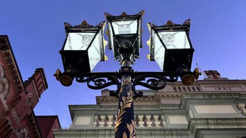 The photo is taken from below the street lamp. It is black with gold leaves engraved on it. The lamp has three lights at the top of it.