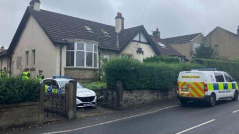 Police officers stand outside the house where the man was found, with a police van and car also parked outside