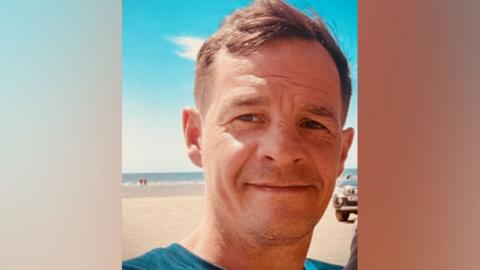 Selfie of Craig Foy standing in the sunshine on a beach with blue sky. He is smiling, has shot dark hair and is wearing a teal blue t-shirt