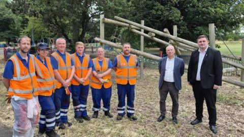 Community caretakers Liam Price, Simone Widdowson, Jonathan Bannister, Alex Tarling, Samantha Wilson and Karl Carr with Executive Member for Environment and Sustainability Councillor Peter Gavigan and Mayor Chris Cooke at Pallister Park. The community caretakers are wearing blue shirts and trousers, with orange vests. Peter Gavigan and Chris Cooke are wearing trousers, white shirts and jackets.