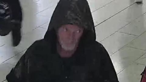 Still from a CCTV camera of a man in a shopping centre that police want to speak to. He has a grey beard and is wearing a black coat with a hood.