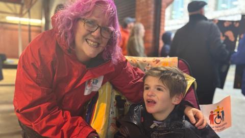 A woman with purple hair and dark rimmed glasses and a red coat leans over a boy in a black rain coat in a wheelchair