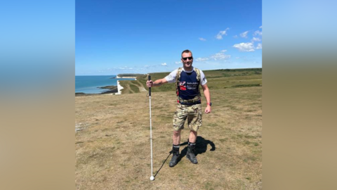 A man wearing camouflage shorts and holding a walking cane. He is stood on a cliff with blue skies in the background.