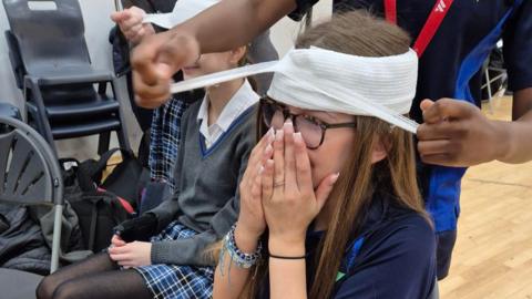 A girl, with her head wrapped in a bandage. She has her hands up to her face, and is wearing glasses. Other pupils are sat behind her and someone is behind her. Chairs are to the left. 