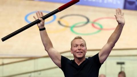 Chris Hoy, wearing a black t-shirt, holds a baton in the air during a ceremony at a velodrome in Paris. Behind him is the velodrome with the five Olympic rings