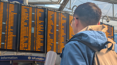 A man with short black hair and wearing glasses, a blue jacket and a beige bag is stood in Manchester Piccadilly station looking at the departure board.