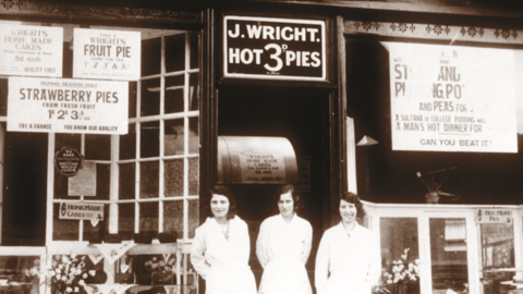 A black and white photo shows three women stood outside a shop with a sign that says "J Wright. Hot pies 3d". There are other adverts for food in the window either side of the women. They have their hair tired back and are wearing a light-coloured uniform.
