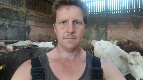 A man with brown hair in a cow shed surrounded by cows