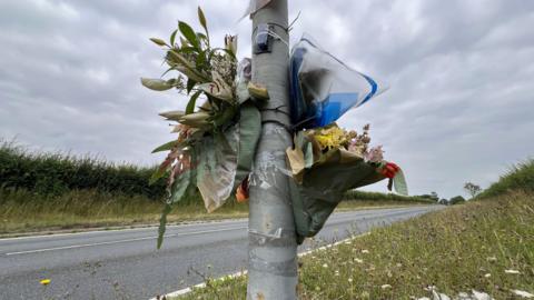 Several floral tributes, tied to a grey lampost, next to the side of a main road. There is a green verge and hedgerow on both sides of the road, and dark grey clouds in the sky above.