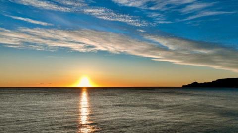 Sun rising above the horizon, reflected onto a dark coloured sea, cliff to the right under a light blue sky with streaky cloud