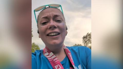 Micky Marsden with brown hair tied back and green sunglasses resting on her head wearing a blue t-shirt with the PAC charity logo and the words Positive Action in the Community. She is at the finishing line of the London Marathon with the medal around her neck. She is smiling.