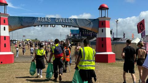 People walking around the Boardmasters festival site. Some are wearing yellow high-vis jackets and are picking up litter.