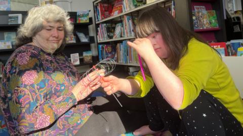 Susan Arthur and Molly use a sensory kit. Molly, who has Down's syndrome, has long brown hair. She is dressed in a green jumper with black trousers which have small silver stars. Molly is holding a spinning wheel and a piece of purple tubing. Susan is sat next to her, with short curly fair hair and wearing a colourful patterned top.