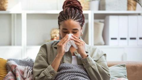 A young woman sits on her sofa, blowing her nose into a tissue