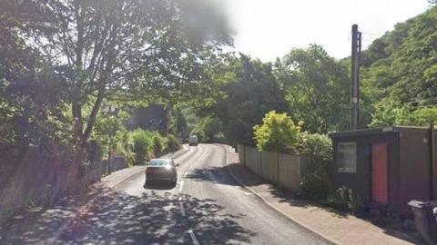 A car travels along a narrow rural road on a sunny day. The road is flanked by green-leafed trees all around and a brown shed with a red door sits on the right-hand side of the picture. 