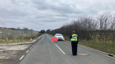 A police officer in high visibility jacket with her back to the camera stands in the middle of the road in front of a police car with its lights on and a red sign saying road closed