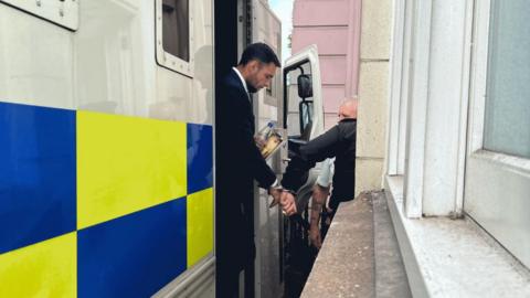 Dylan Pounds being taken out of a police van outside court. He is handcuffed to a prison officer. He is wearing a suit and white shirt and is carrying a bottle of water and a book. 