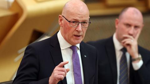 A bald man wearing glasses, a black jacket and white shirt with purple tie raises a finger as he answers a question in the Parliament chamber
