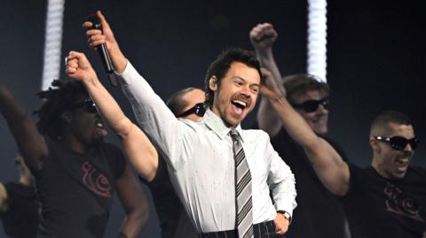 Harry Styles performs at the Brit Awards. He is wearing a grey office shirt and tie combination.