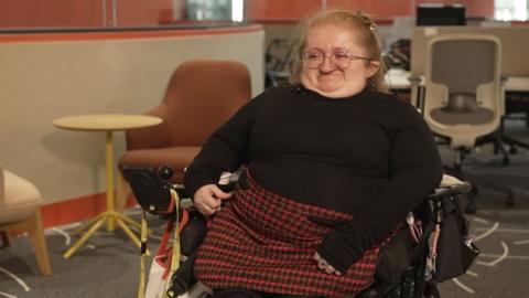 A woman in a wheelchair. She is wearing a black jumper and a white and black skirt.