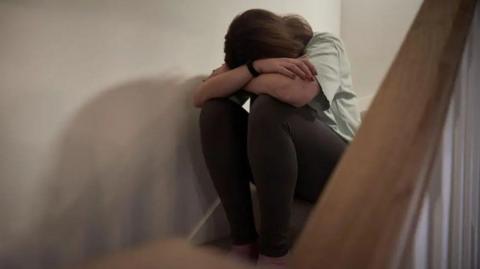 A woman sits on a staircase with her head resting on her crossed arms. She is wearing dark leggings and a light-coloured T-shirt. 