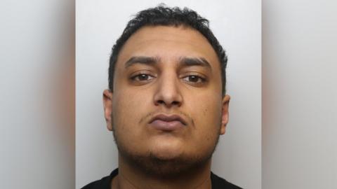 A close up crop of a mugshot. Janjua has brown eyes, brown hair, a thin moustache and thin beard.
