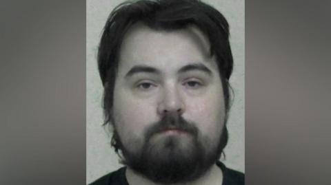 Mugshot of McLean. He has large head with very pale skin and scruffy long black hair and beard.