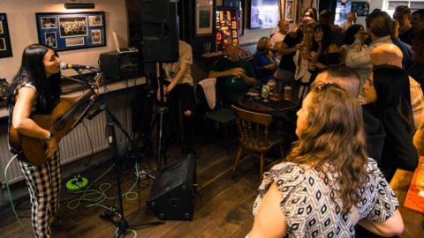 A woman with a guitar sings in front of a crowd in a pub. She is holding a guitar and singing into a microphone. Next to her is a speaker. People are standing beside the bar and around small pub tables to watch her. 