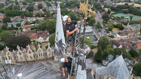 Two men in black tops and shorts, in harnesses and with white helmets are standing at the top of a ladder looking at a golden weather metal vane at the top of a cathedral's pinnacle. Below them is a city with buildings and trees.