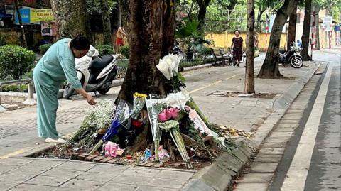 A woman in mint green clothes bends over a ring of flower bouquets arranged around the trunk of a roadside tree