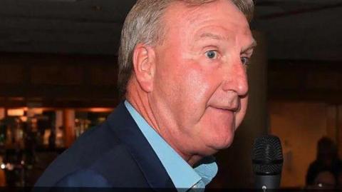 A side view of a man who has grey hair and is wearing a dark blue suit jacket and a light blue shirt. 