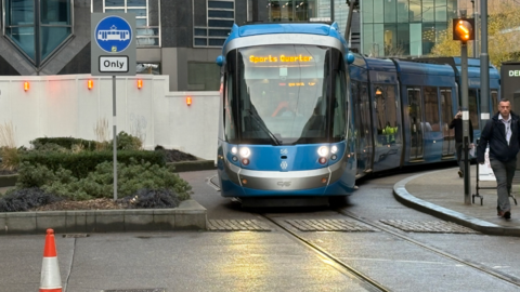 A blue tram with an orange sign for Sports Quarter trundles through an area of the Birmingham city centre. A man walks along the street to the right of the tram. The road is wet and an orange cone can be seen on the left of the picture.