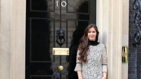 Zoe Clark-Coates is standing outside the black door of No.10 Downing Street. She has long dark hair and is smiling. 