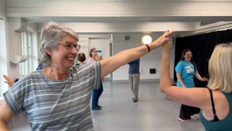 A woman with grey shoulder length hair and a striped top has her left arm outstretched and raised dancing with her partner, whose face isn't visible. A handful of other dancers and an instructor is visible in the background. 