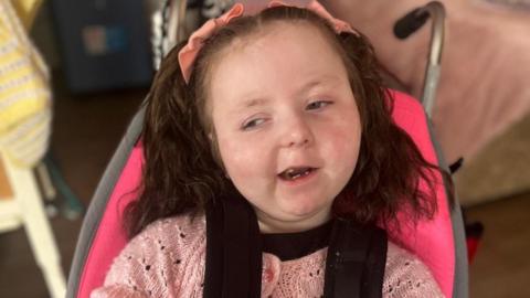 A girl, around 8 years old, with pig tails in her hair, sat in a chair. She is wearing a pink woollen top. 