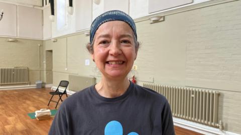 Photo of Massey, who is looking into the camera and smiling. She is wearing a blue headscarf with yellow stripes on it, with her curly brown hair just visible underneath it. Massey is wearing a dark grey t-shirt with a large light blue flower motif just visible. She is stood inside a hall, with has light green and white painted walls and a wooden floor. Behind Massey is a black fold-away chair and a green yoga mat.