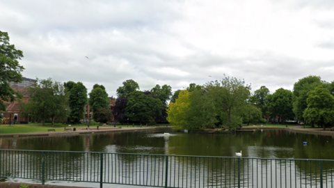A parkland surrounding a large lake. Trees can be seen in the background with fencing on one side of the water in the foreground 