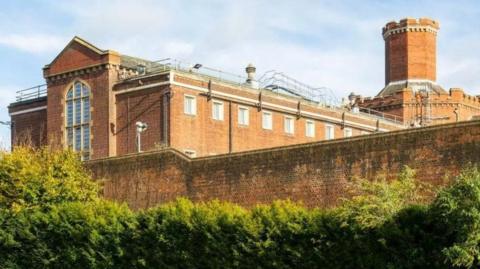 Red brick wall surrounds large building with long glass window and end, multiple windows at height and a turret