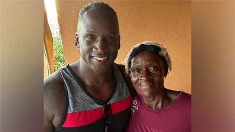 A man with short black hair is standing and smiling with his arm around a woman. He is wearing a grey, black and red striped vest. The woman has short blonde hair and a black headband and is wearing a dark pink top.